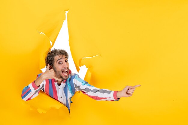 Sonriente y emocional joven posa en fondo de agujero de papel amarillo rasgado apuntando algo y haciendo gesto de llamarme