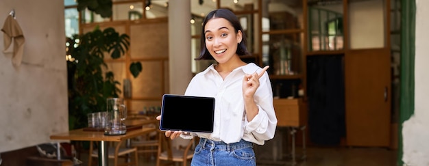 Foto gratuita sonriente empresaria asiática demostrando mostrar la pantalla de la tableta y apuntando a pancartas publicitarias