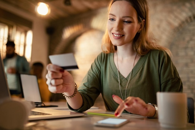 Foto gratuita sonriente empresaria haciendo banca electrónica con tarjeta de crédito y teléfono móvil mientras trabaja hasta tarde en la oficina