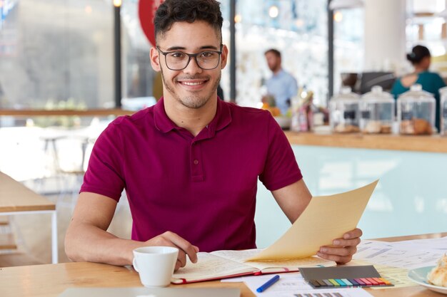 Sonriente empresario datisfied sostiene papeles, viste ropa informal, se prepara para el taller de capacitación, lee la información necesaria, analiza la documentación, posa contra el interior del café. Las condiciones de trabajo