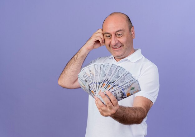 Sonriente empresario maduro casual estirándose y mirando dinero y tocando la cabeza aislada sobre fondo púrpura con espacio de copia