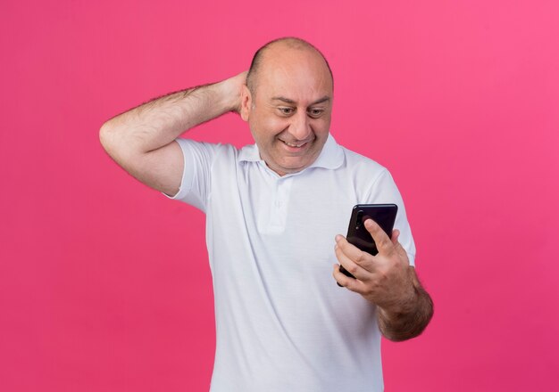 Sonriente empresario maduro casual sosteniendo y mirando el teléfono móvil manteniendo la mano detrás de la cabeza aislada sobre fondo rosa con espacio de copia