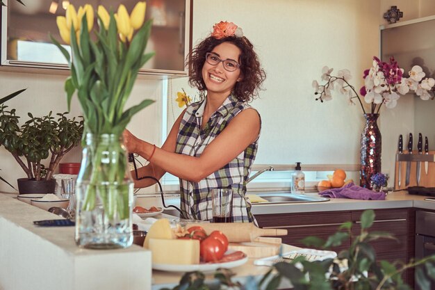 Sonriente encantadora chica hispana rizada cocinando en su cocina. Mezcla los ingredientes con una batidora de mano.