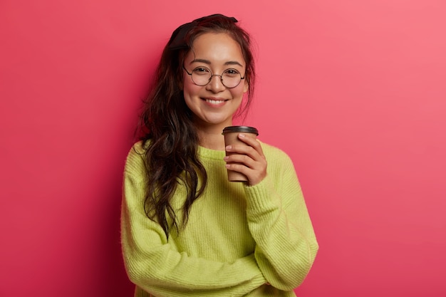 Sonriente y encantadora modelo femenina disfruta tomando café durante el tiempo libre, tiene buen humor, habla informal con el intelocutor, mira directamente a la cámara