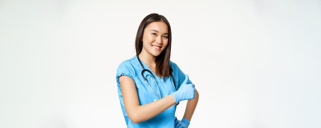 Sonriente enfermera médica asiática en matorrales muestra vacunas contra la gripe de la mano o la campaña de vacunación covid19 mostrando los pulgares hacia arriba fondo blanco