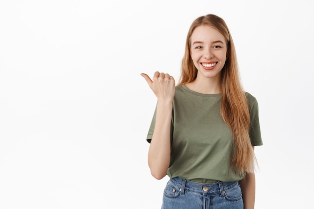 Sonriente estudiante confiada apuntando con el pulgar hacia la izquierda y mirando determinada, dar consejos, recomendar este producto, mostrando publicidad en el espacio de la copia, pared blanca