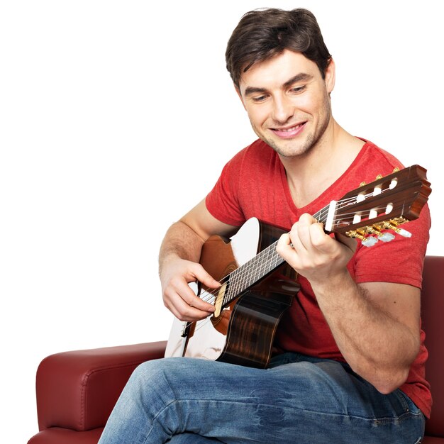 Sonriente guitarrista juega en el guitat isolatade acústico sobre fondo blanco. Apuesto joven se sienta con la guitarra en el diván