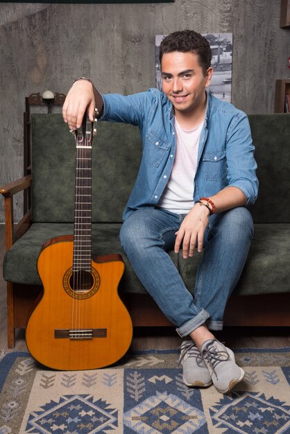 Sonriente guitarrista sosteniendo una hermosa guitarra y sentado en el sofá. Foto de alta calidad