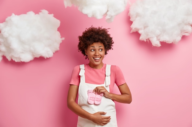 Sonriente y hermosa joven se regocija de su embarazo, feliz de tener un bebé, sostiene calcetines pequeños sobre la barriga, usa ropa casual para embarazadas, mira a un lado, se para en el interior. Concepto de maternidad feliz