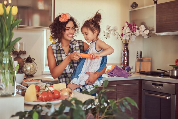 Sonriente y hermosa madre hispana rizada le enseña a su linda hijita a preparar pizza en la cocina.