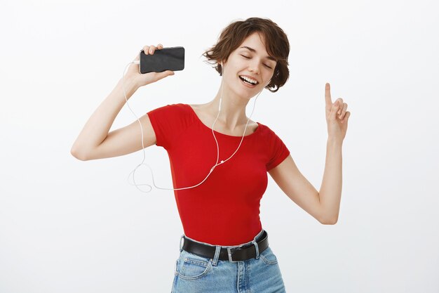 Sonriente a hermosa mujer morena bailando despreocupada, escuchando música y bailando en auriculares, sosteniendo el teléfono inteligente