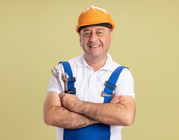 Sonriente hombre adulto constructor en uniforme se encuentra con los brazos cruzados sosteniendo una llave y una llave inglesa aislada en la pared verde oliva