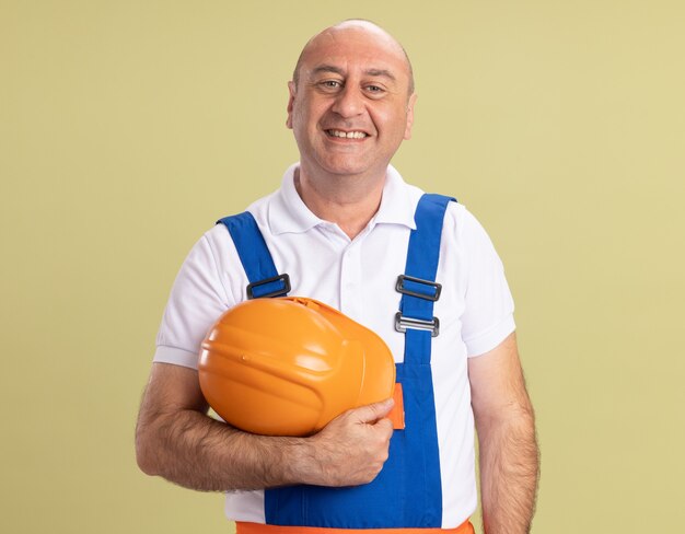Sonriente hombre adulto constructor en uniforme tiene casco de seguridad aislado en la pared verde oliva