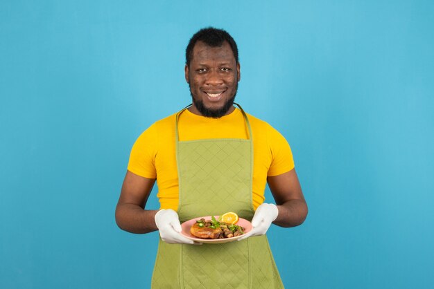 Sonriente hombre afroamericano con barba vistiendo delantal con un plato de comida en la mano, se encuentra sobre la pared azul.