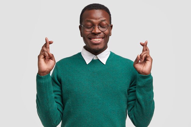 Sonriente hombre afroamericano reza por el cumplimiento de su deseo, cruza los dedos, desea buena suerte