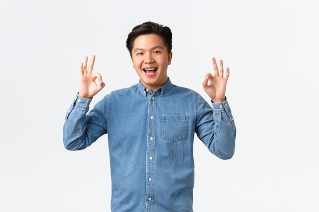 Sonriente hombre asiático satisfecho con tirantes en camisa azul, mostrando un gesto bien, felicitando a la persona con un trabajo excelente, bien hecho, recomiendo un servicio perfecto o calidad, pared blanca