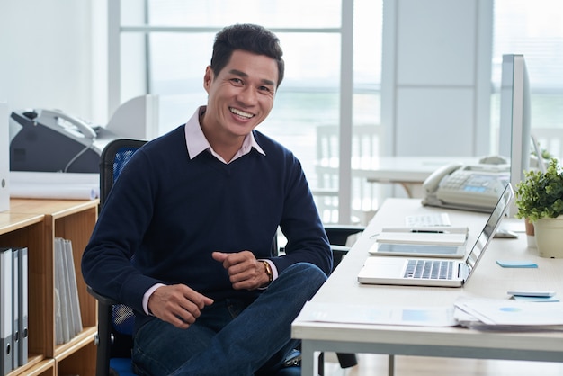 Sonriente hombre asiático sentado en el escritorio frente a la computadora portátil en la oficina y mirando a la cámara