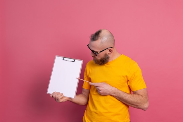 Sonriente hombre barbudo con anteojos vistiendo ropa casual sosteniendo un portapapeles con un papel en blanco apuntando