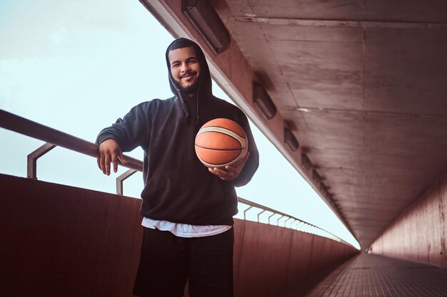 Sonriente hombre barbudo de piel oscura vestido con una capucha negra y pantalones cortos deportivos apoyado en una barandilla mientras está de pie con baloncesto, mirando una cámara.