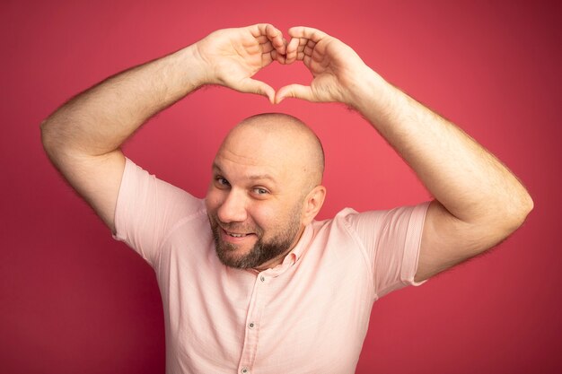 Sonriente hombre calvo de mediana edad con camiseta rosa levantando gesto de corazón aislado en rosa