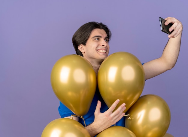 Foto gratuita sonriente hombre caucásico guapo se encuentra con globos de helio sosteniendo y mirando el teléfono tomando selfie