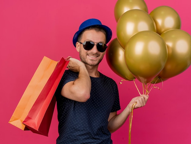 Sonriente hombre caucásico guapo en gafas de sol con sombrero de fiesta azul sostiene globos de helio y bolsas de papel aisladas sobre fondo rosa con espacio de copia