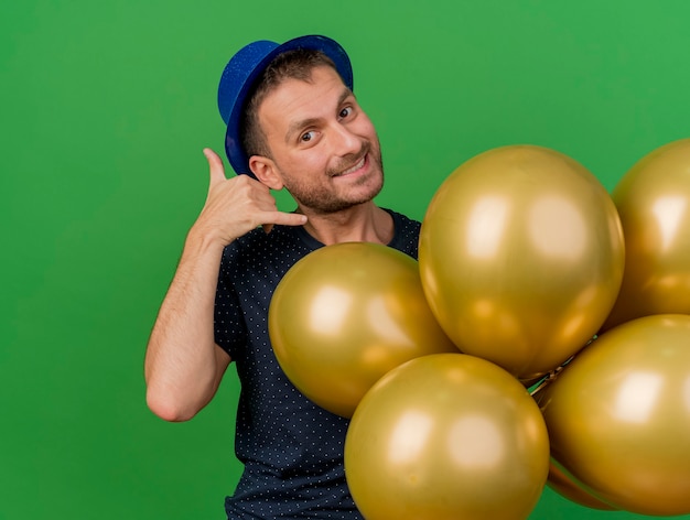 Sonriente hombre caucásico guapo con gestos de sombrero de fiesta azul me llaman firmar y sostiene globos de helio aislados sobre fondo verde con espacio de copia