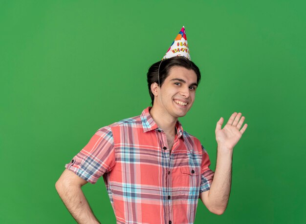 Sonriente hombre caucásico guapo con gorro de cumpleaños se encuentra con la mano levantada