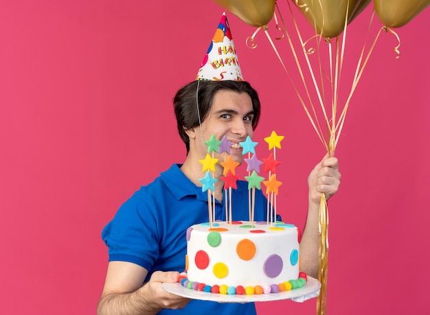 Sonriente hombre caucásico guapo con gorro de cumpleaños sostiene globos de helio y pastel de cumpleaños