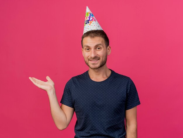 Sonriente hombre caucásico guapo con gorro de cumpleaños sostiene la mano abierta aislada sobre fondo rosa con espacio de copia