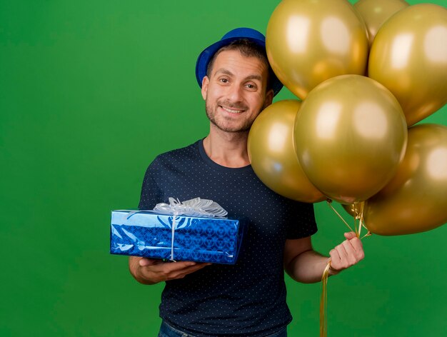 Sonriente hombre caucásico guapo con gorro de fiesta tiene globos de helio y caja de regalo aislado sobre fondo verde con espacio de copia