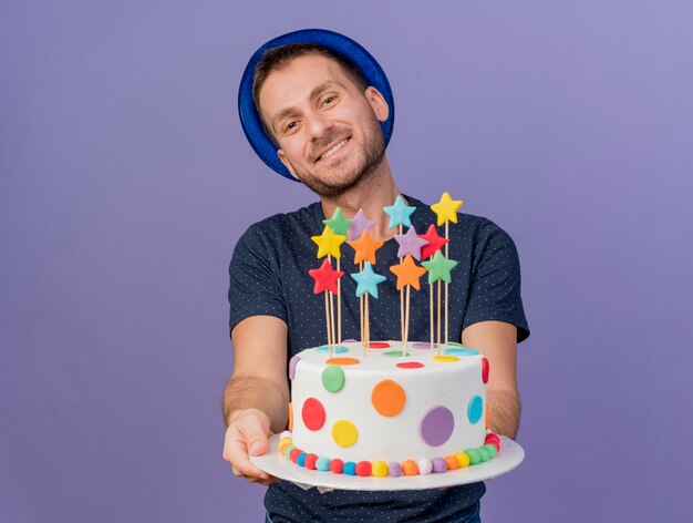 Sonriente hombre caucásico guapo con sombrero azul sostiene la torta de cumpleaños mirando a cámara aislada sobre fondo púrpura con espacio de copia