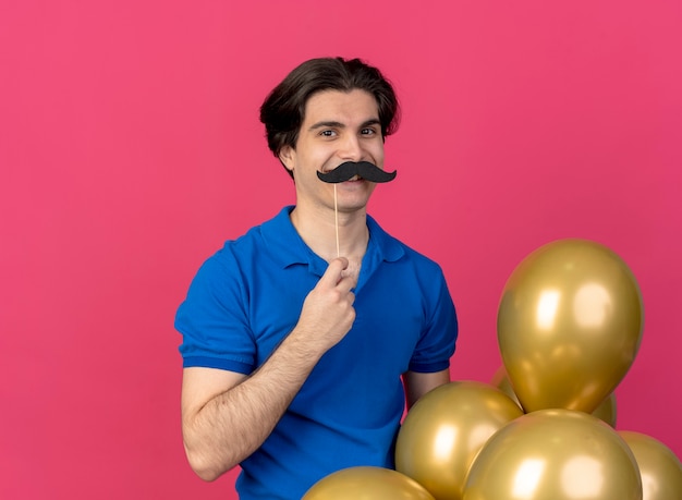 Sonriente hombre caucásico guapo con sombrero de fiesta azul sostiene globos de helio y bigote falso en el palillo