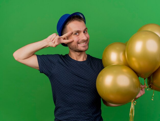 Sonriente hombre caucásico guapo con sombrero de fiesta azul sostiene globos de helio y gestos signo de mano de victoria aislado sobre fondo verde con espacio de copia