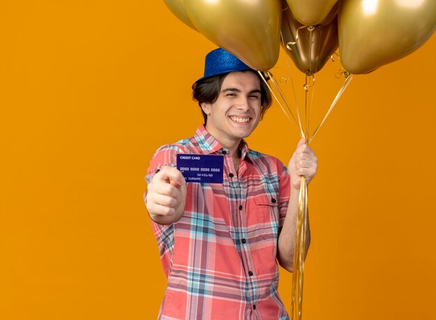 Sonriente hombre caucásico guapo con sombrero de fiesta azul tiene globos de helio y tarjeta de crédito