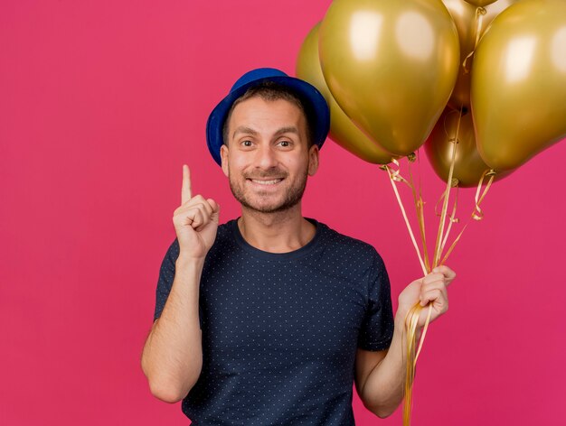 Sonriente hombre caucásico guapo vestido con gorro de fiesta azul sostiene globos de helio apuntando hacia arriba aislado sobre fondo rosa con espacio de copia