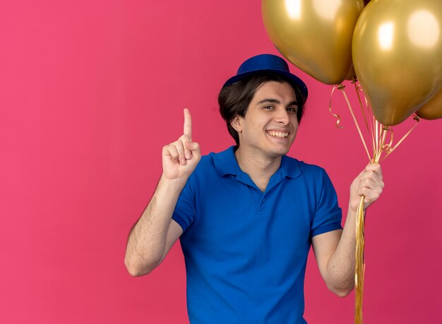 Sonriente hombre caucásico guapo vestido con gorro de fiesta azul sostiene globos de helio apuntando hacia arriba