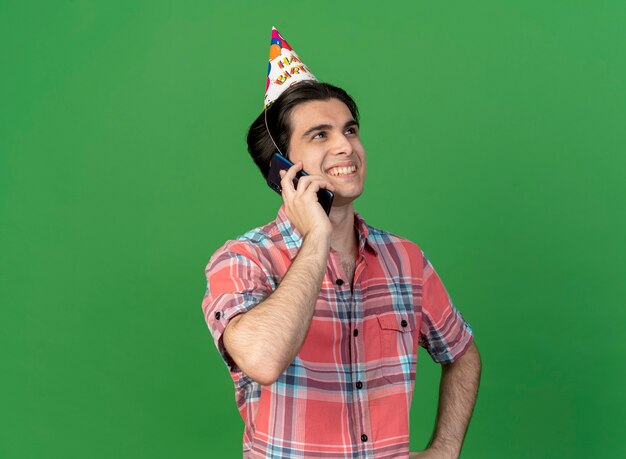 Sonriente hombre caucásico guapo vistiendo gorro de cumpleaños habla por teléfono