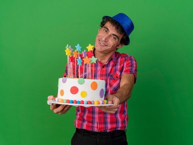 Sonriente hombre caucásico de mediana edad con gorro de fiesta mirando a la cámara estirando el pastel de cumpleaños hacia la cámara aislada sobre fondo verde con espacio de copia