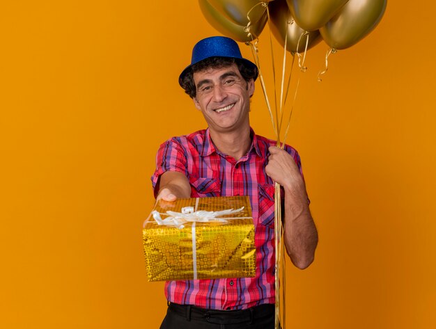 Sonriente hombre caucásico de mediana edad con gorro de fiesta sosteniendo globos mirando a la cámara estirando el paquete de regalo hacia la cámara aislada sobre fondo naranja con espacio de copia