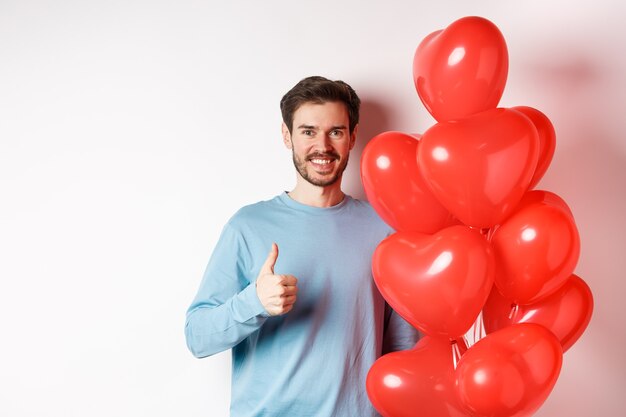 Sonriente hombre caucásico de pie con globo de corazón, preparar sorpresa para el amante en el día de San Valentín, mostrando los pulgares hacia arriba y mirando a cámara, fondo blanco.