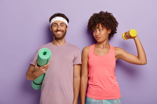 Sonriente hombre complacido posa con una colchoneta de fitness, viste una camiseta morada y una diadema, una encantadora mujer deportiva mira a su marido, entrena bíceps con peso, párese hombro con hombro en interiores. Aeróbicos y personas