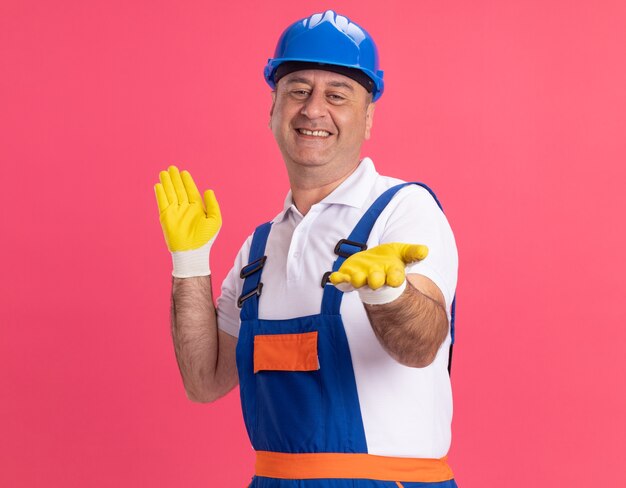 Sonriente hombre constructor adulto en uniforme con guantes protectores tiene las manos abiertas aisladas en la pared rosa