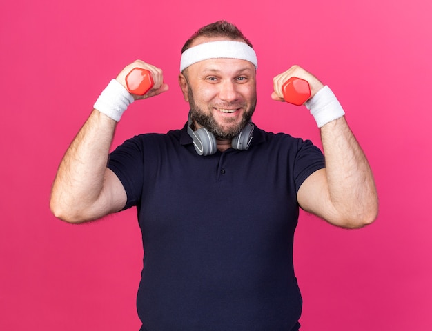Sonriente hombre deportivo eslavo adulto con auriculares con diadema y muñequeras sosteniendo pesas aislado en la pared rosa con espacio de copia