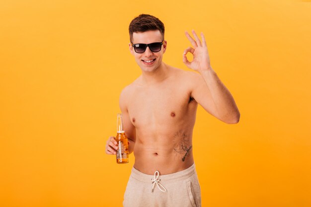 Sonriente hombre desnudo en pantalones cortos y gafas de sol sosteniendo una botella de cerveza y mostrando un signo bien sobre amarillo