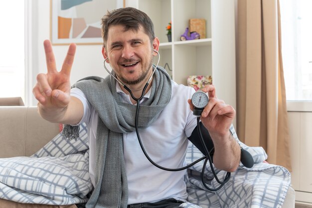 Sonriente hombre enfermo con bufanda alrededor del cuello midiendo su presión con esfigmomanómetro y gesticulando el signo de la victoria sentado en el sofá de la sala de estar