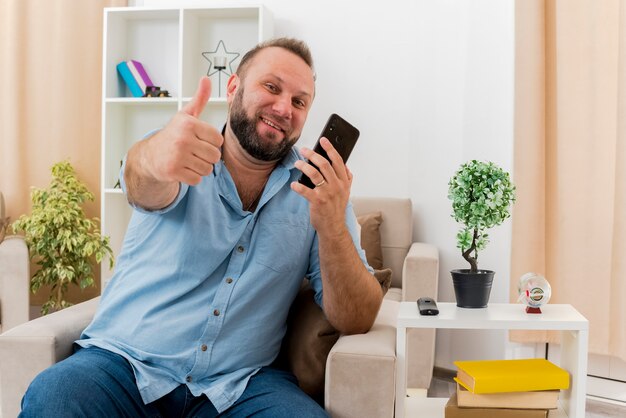 Sonriente hombre eslavo adulto se sienta en el sillón Thumbs up sosteniendo el teléfono dentro de la sala de estar