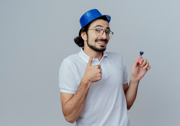 Sonriente hombre guapo con gafas y sombrero azul sosteniendo silbato con el pulgar hacia arriba aislado en blanco