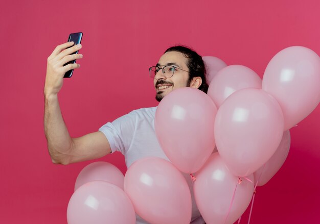 Sonriente hombre guapo con gafas sosteniendo globos y tomar un selfie aislado en rosa