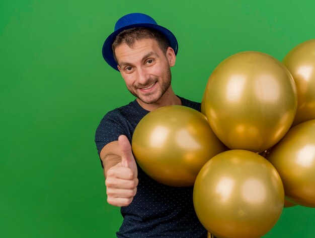 Sonriente hombre guapo con gorro de fiesta azul sostiene globos de helio y pulgares arriba aislados en la pared verde con espacio de copia